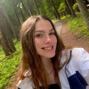 A college student with long brown hair smiles at the camera in a selfie taken along a forested path.